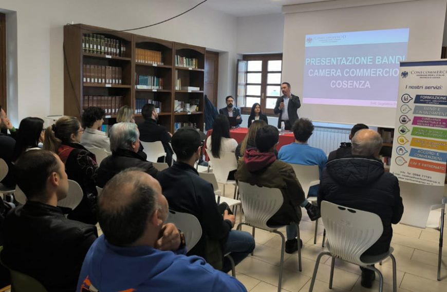 Un evento di presentazione dei bandi della Camera di Commercio di Cosenza, organizzato da Confcommercio. La sala è piena di partecipanti seduti, mentre un relatore parla davanti a un pannello con il titolo della presentazione. Un roll-up di Confcommercio elenca i servizi offerti.